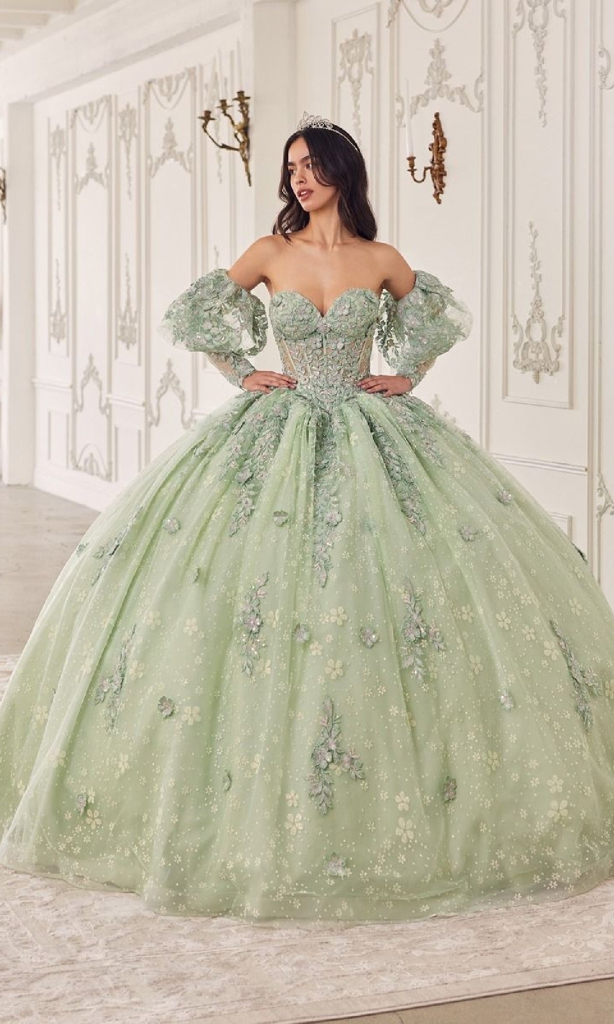 A woman wears the Quinceanera Dress 15722 by Ladivine—a light green gown with floral embellishments and removable puff sleeves—while standing in an ornate, white room.